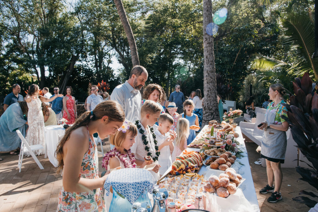 A group of people, including children, are gathered outdoors at a buffet table filled with various foods. Theyre surrounded by trees and sunlight filters through the foliage, creating a festive atmosphere. Everyone appears to be enjoying the gathering at a brunch wedding private chef booking on Maui.