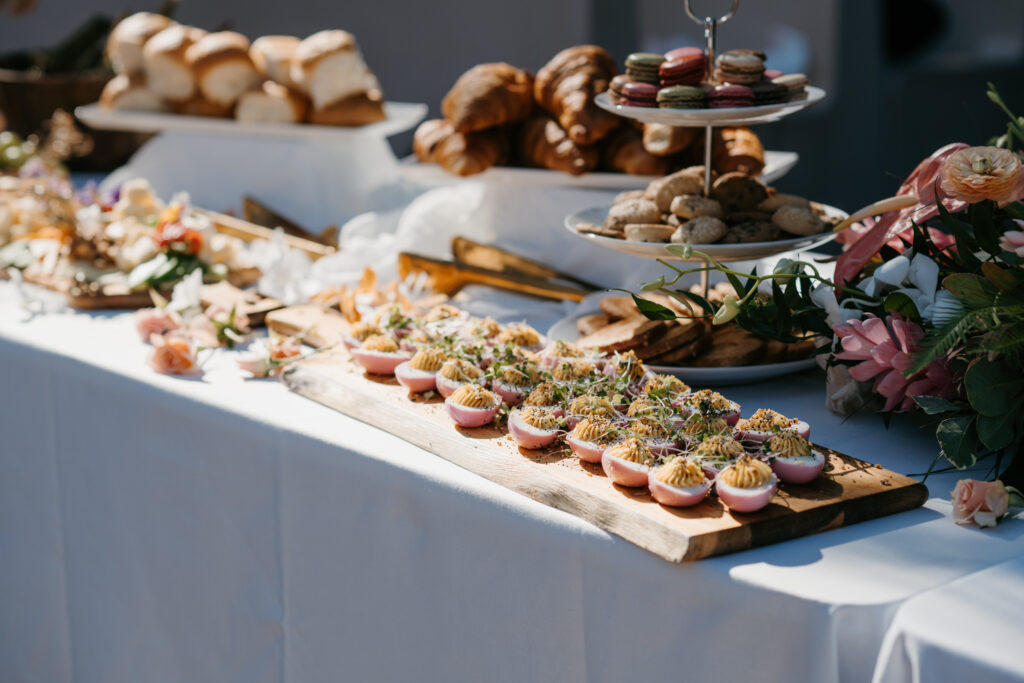 A table set for a Maui High Tea or a styled grazing board features a variety of baked goods, including cupcakes with pink frosting on a wooden board, croissants on trays, and assorted macarons on a tiered stand. Decorative flowers are arranged around the table to enhance the tropical elegance.