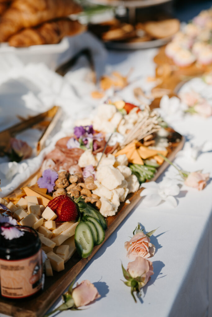 A charcuterie board on a white table with cheese, nuts, sliced cucumber, strawberries, and crackers, garnished with flowers brunch wedding private chef booking on Maui. In the background, croissants and pastries are visible. Scattered pink roses decorate the table.