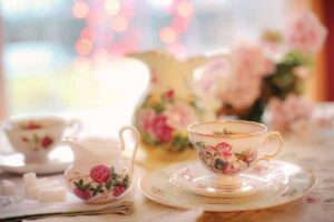 A delicate tea set with floral patterns, perfect for Maui High Tea, featuring a cup and saucer, sugar cubes, and a small creamer. The background is softly blurred with pink and white flowers reminiscent of Mauis lush scenery, creating a warm, inviting atmosphere.