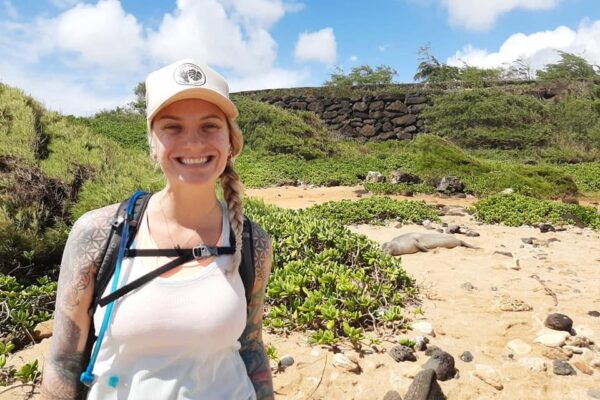 A smiling woman with a tattooed arm and a cap stands on a sandy beach with greenery; behind her, a monk seal is resting on the sand under a sunny sky. Erin Elizabeth captures the serene moment beautifully.