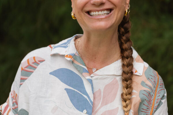 A woman with a long braided ponytail smiles widely. She wears a colorful patterned shirt and gold hoop earrings, reminiscent of Erin Elizabeth’s vibrant style. Green foliage is blurred in the background.