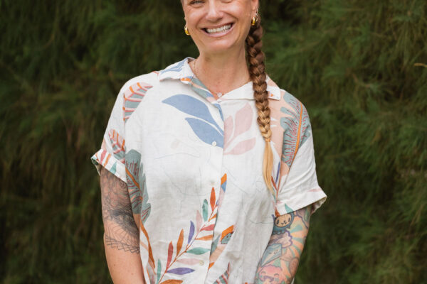 A smiling person with a long braid, tattoos, and a short-sleeved patterned shirt stands in front of lush green foliage, hands clasped together—capturing the vibrant spirit of Erin Elizabeth.