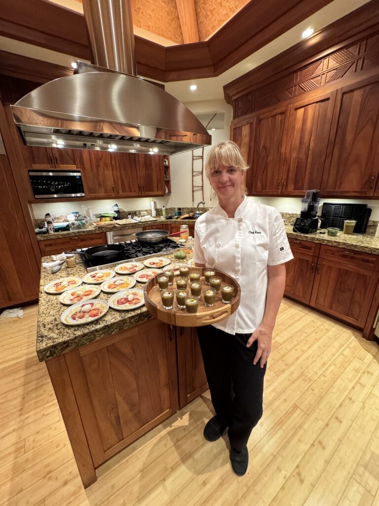 Chef Kena Holtine, dressed in a white coat, stands in a wooden kitchen holding a tray of small green appetizers. Plates with various dishes are arranged on the granite countertop behind her.