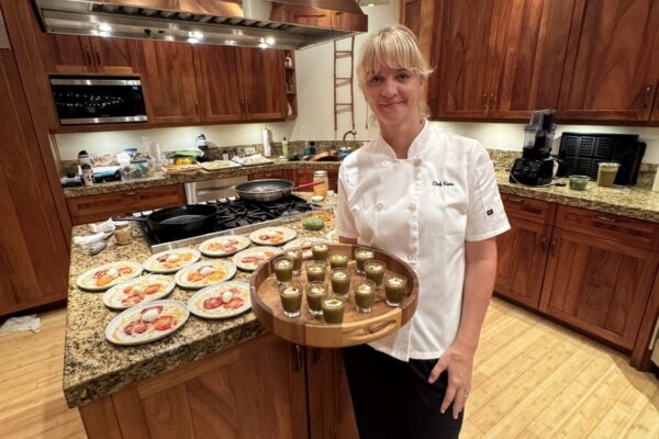 Chef Kena Holtine, dressed in a white coat, stands in a wooden kitchen holding a tray of small green appetizers. Plates with various dishes are arranged on the granite countertop behind her.
