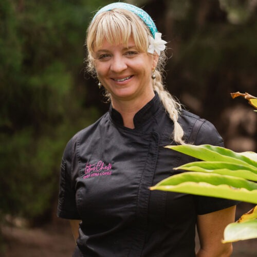 Kena Holtine, wearing a black chef’s coat and a blue headband adorned with a white flower, stands outside smiling, with green foliage in the background.