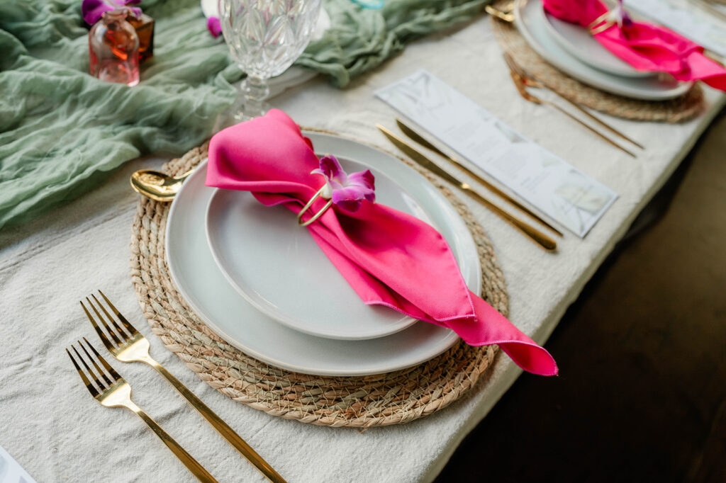 A table setting inspired by Chef Kyra Mirian Bramble features white plates on a woven placemat, gold utensils, a pink napkin with a purple flower, and a crystal glass atop a white cloth with a green fabric runner.