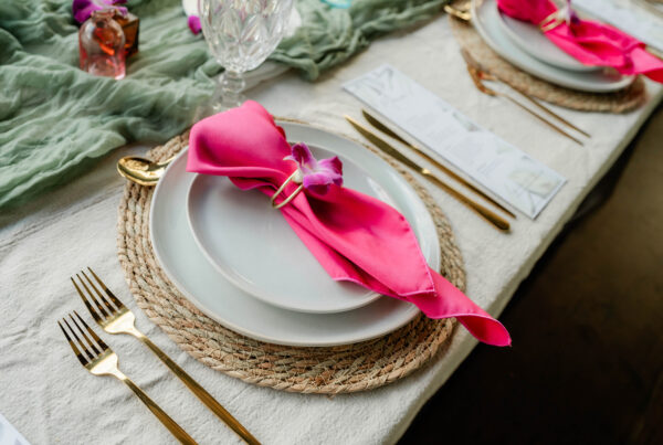 A table setting inspired by Chef Kyra Mirian Bramble features white plates on a woven placemat, gold utensils, a pink napkin with a purple flower, and a crystal glass atop a white cloth with a green fabric runner.