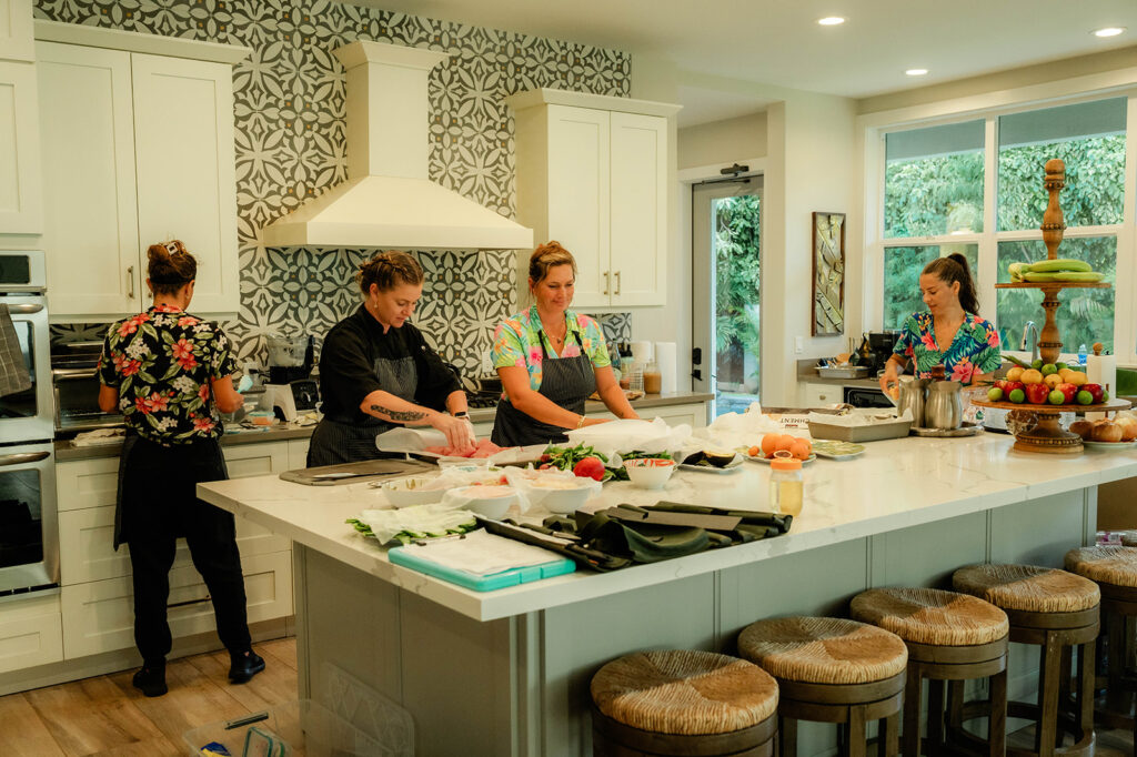 Chef Kyra Mirian Bramble leads four people preparing food in a bright, modern kitchen with a patterned backsplash. Various ingredients and utensils cover the large island, while three stools sit in the foreground amid the busy yet collaborative atmosphere.