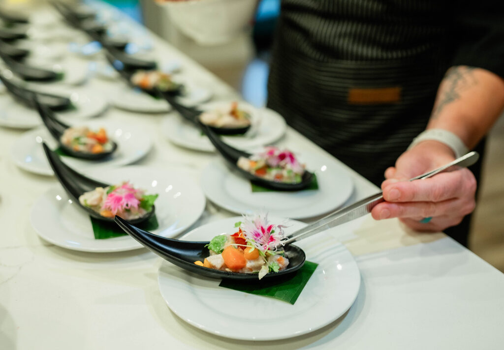 Chef Kyra Mirian Bramble arranges colorful, flower-garnished appetizers on black curved spoons placed on white plates in a row on a counter.