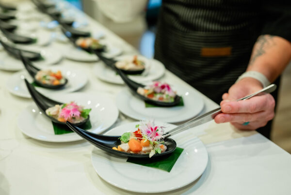 Chef Kyra Mirian Bramble arranges colorful, flower-garnished appetizers on black curved spoons placed on white plates in a row on a counter.