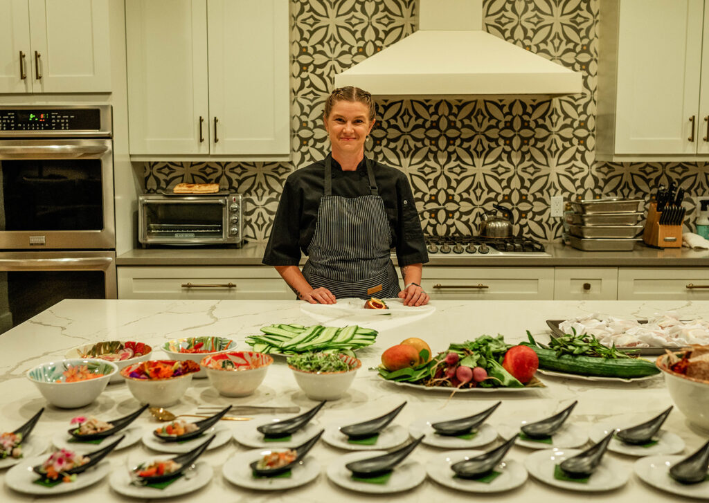 Chef Kyra Mirian Bramble, dressed in a black uniform, stands in a modern kitchen behind a marble counter filled with fresh vegetables, neatly arranged ingredients, and prepared dishes on black spoons. White cabinets and patterned tile backsplash complete the scene.
