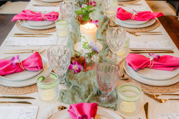 A long dining table set with white plates, crystal glasses, gold cutlery, and bright pink napkins by Chef Kyra Mirian Bramble. The centerpiece features candles and flowers, with greenery running down the middle. Black chairs surround the table.