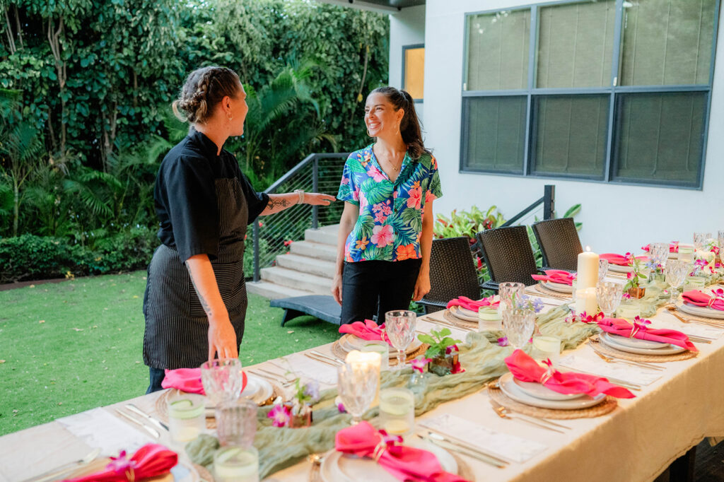 Two women stand and smile at each other beside a long, outdoor dining table set with pink napkins, plates, glasses, and candles. Lush greenery and a modern house serve as the backdrop for Chef Kyra Mirian Bramble’s beautifully curated setting.