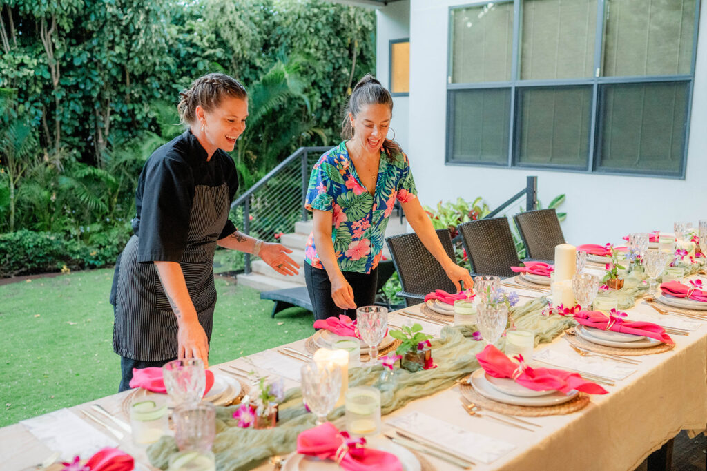 Two women smile as they set a long outdoor dining table decorated with pink napkins, candles, glassware, and floral arrangements in a lush garden setting curated by Chef Kyra Mirian Bramble.