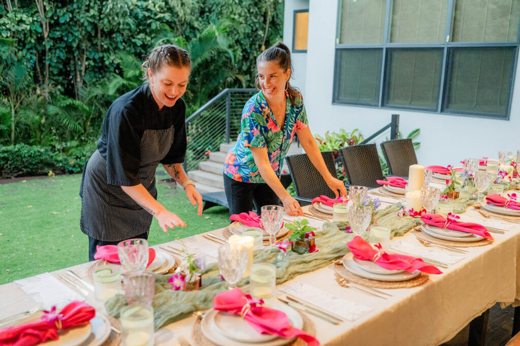 Chef Kyra Mirian Bramble, in a black chef uniform, and a woman in a colorful shirt arrange a beautifully set outdoor dining table with pink napkins, candles, and floral decorations in a garden setting.