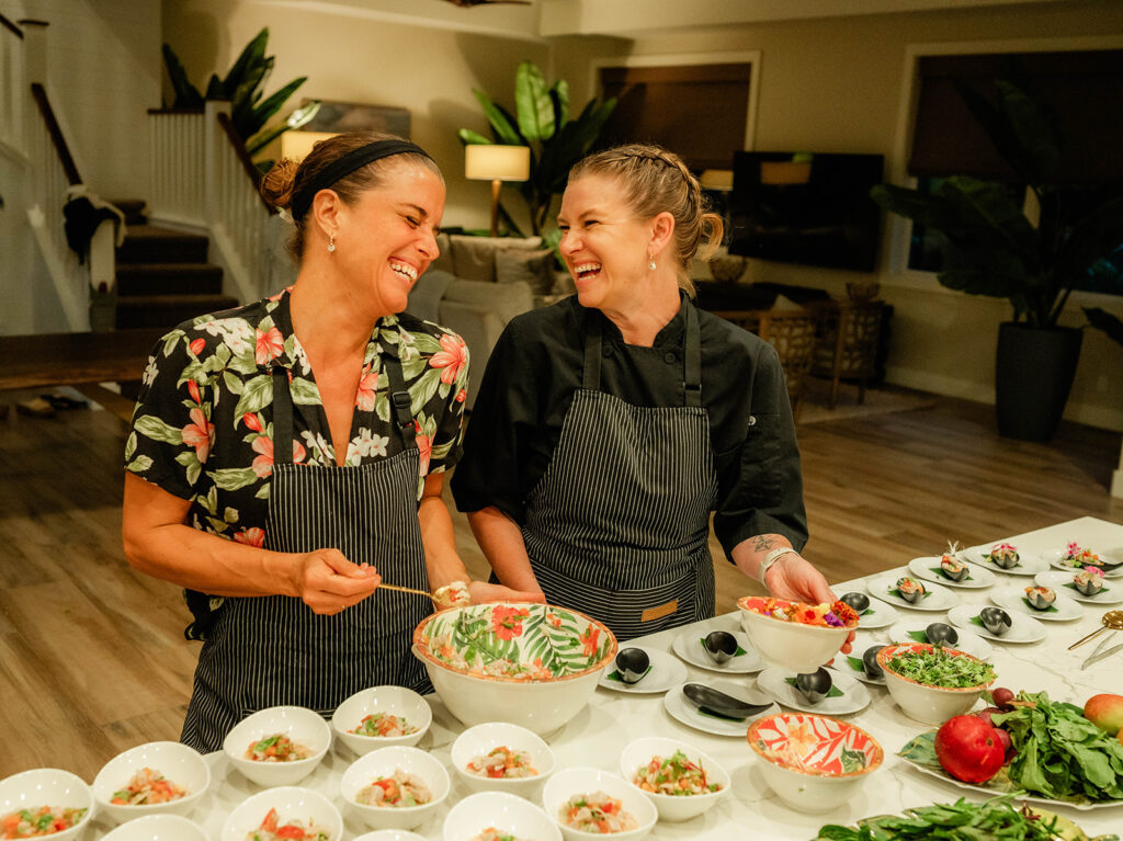 Chef Kyra Mirian Bramble and a friend, both wearing aprons, smile and laugh together behind a counter filled with vibrant salads and bowls of colorful food in a bright, modern kitchen setting.
