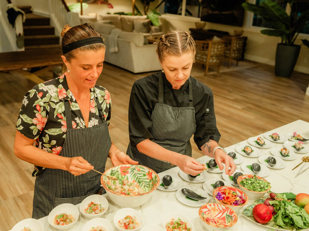 Two women in aprons, including Chef Kyra Mirian Bramble, prepare and arrange colorful bowls of food on a kitchen counter, surrounded by fresh ingredients and herbs, in a bright, modern kitchen.