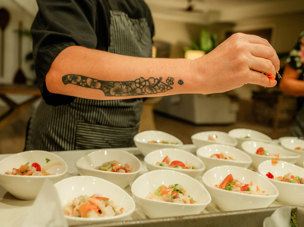 Chef Kyra Mirian Bramble, with a floral tattoo on her arm, is garnishing bowls of food—possibly ceviche—in a kitchen. Multiple white bowls filled with colorful ingredients are neatly arranged on the counter.