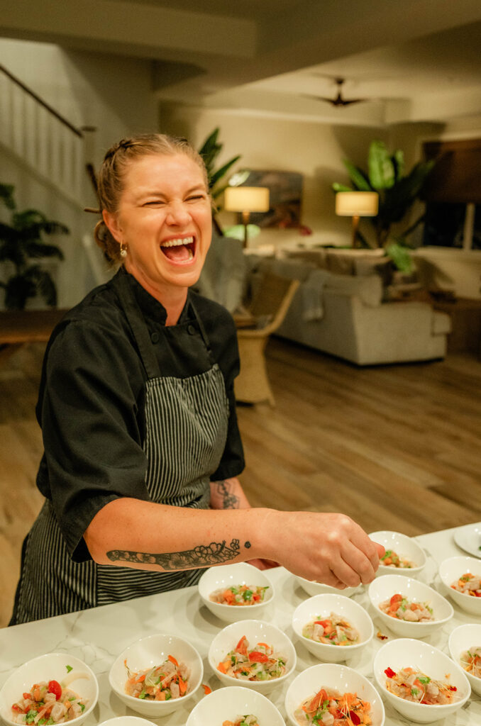 Chef Kyra Mirian Bramble, wearing a black uniform and striped apron, laughs joyfully while preparing several bowls of food on a kitchen counter in a warmly lit, modern home.