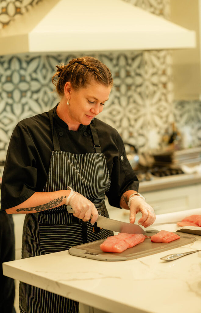 Chef Kyra Mirian Bramble, wearing a black uniform, striped apron, and gloves, slices raw fish on a cutting board in a modern kitchen with patterned tiles in the background.