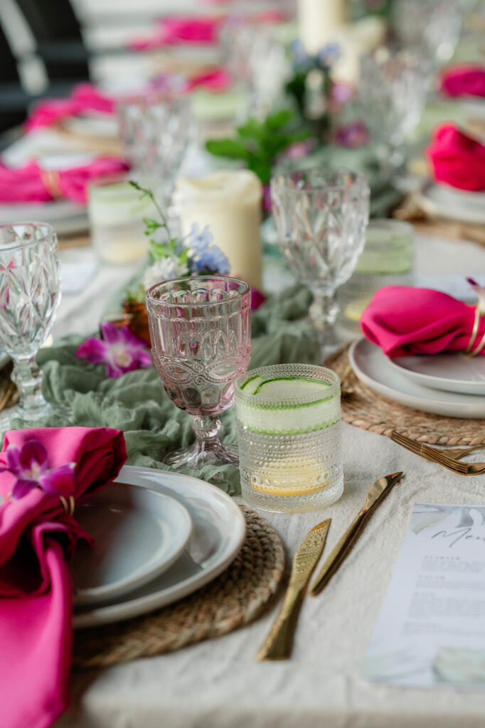 A close-up of a decorative table setting by Chef Kyra with crystal glasses, pink napkins, gold cutlery, and floral accents on a beige tablecloth, creating a vibrant and elegant atmosphere.