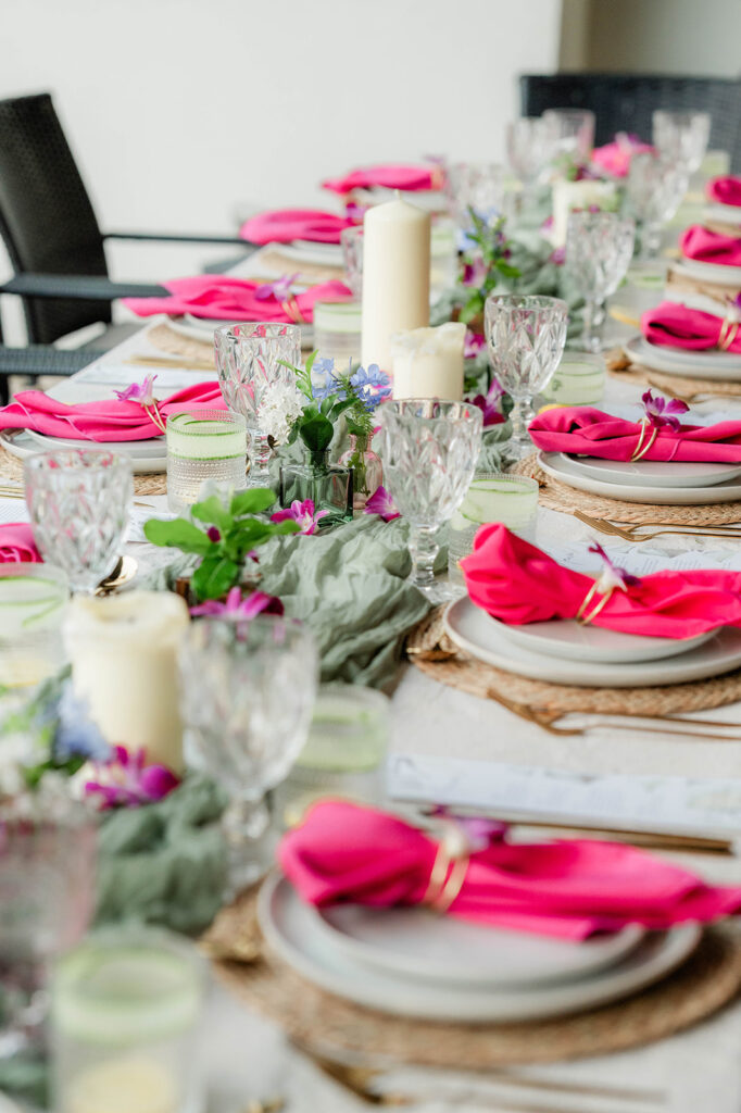 A dining table elegantly set with pink napkins, crystal glasses, white plates, and gold napkin rings. The festive centerpiece features white candles and small purple flowers—a colorful atmosphere inspired by Chef Kyra Mirian Bramble.