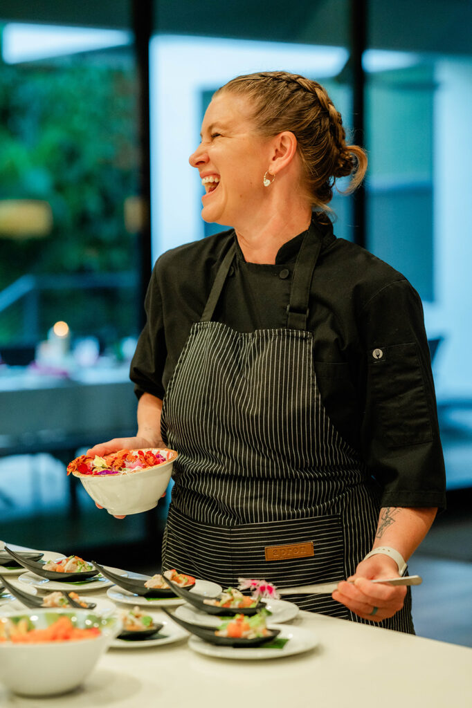 A smiling Chef Kyra Mirian Bramble in a black uniform and striped apron holds a bowl of food, preparing dishes on a counter in a brightly lit, modern kitchen.