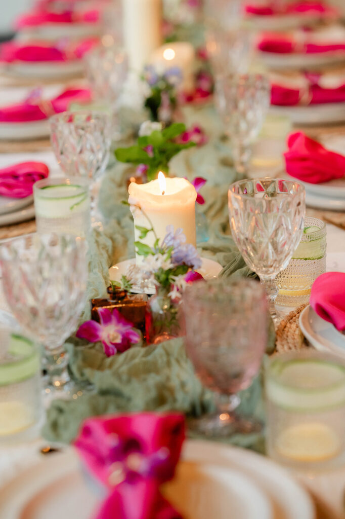 A close-up of an elegant table setting by Chef Kyra Mirian Bramble, featuring crystal glasses, pink napkins, a lit candle, and small flowers along the center. The atmosphere appears festive and inviting.