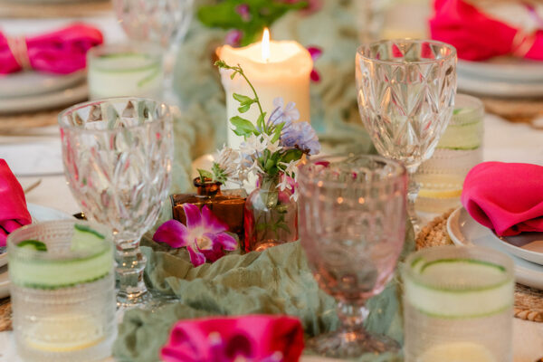 A table set for a meal by Chef Kyra Mirian Bramble features white plates, crystal glasses, green water glasses with cucumber slices, pink napkins, and floral centerpieces. Lit candles and a green table runner add a festive touch.
