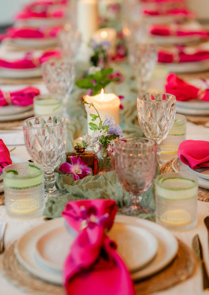 A table set for a meal by Chef Kyra Mirian Bramble features white plates, crystal glasses, green water glasses with cucumber slices, pink napkins, and floral centerpieces. Lit candles and a green table runner add a festive touch.