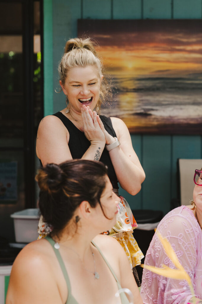 Chef Kyra Mirian Bramble, with a top knot, smiles and clasps her hands in delight while standing behind two seated women at an outdoor gathering, with a sunset beach painting in the background.