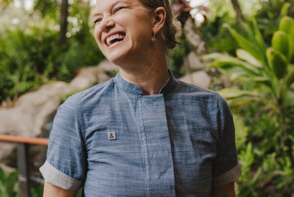 A smiling Chef Kyra Mirian Bramble with tattoos, wearing a blue shirt and black apron, holds a decorated dish on a rectangular plate. Lush green plants and palm trees fill the background, suggesting an outdoor tropical setting.