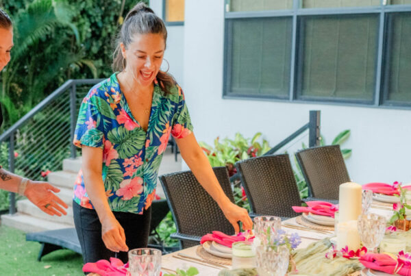 Tara Klitchman and another woman, one in a chef’s uniform and one in a floral shirt, set a long outdoor dining table decorated with pink napkins, candles, and flowers in a garden setting.