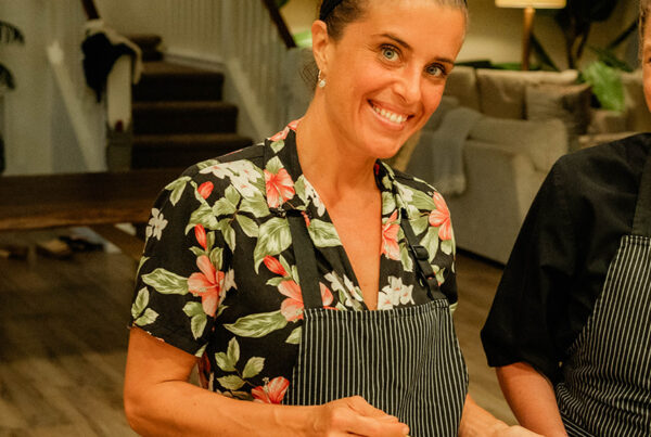 Two smiling women in aprons stand behind a table with several small bowls of colorful food. Angel Green mixes a large bowl while the other stands beside her in a modern, warmly lit kitchen or dining area.