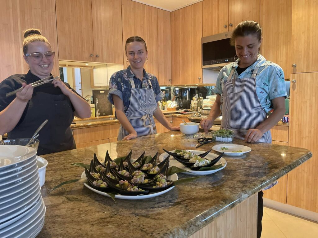 Three people, including Chef Casia Rose, wear aprons and smile in a kitchen around a granite counter topped with neatly plated appetizers. Stacks of plates and cooking ingredients are visible, set against warm wooden cabinets.