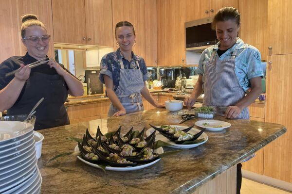 Three people, including Chef Casia Rose, wear aprons and smile in a kitchen around a granite counter topped with neatly plated appetizers. Stacks of plates and cooking ingredients are visible, set against warm wooden cabinets.