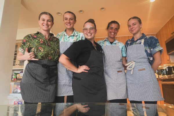 Five people stand indoors, smiling at the camera. Four wear striped aprons over colorful shirts, while Chef Casia Rose, centered in a black apron and glasses, stands out. The setting appears to be a kitchen with warm lighting.
