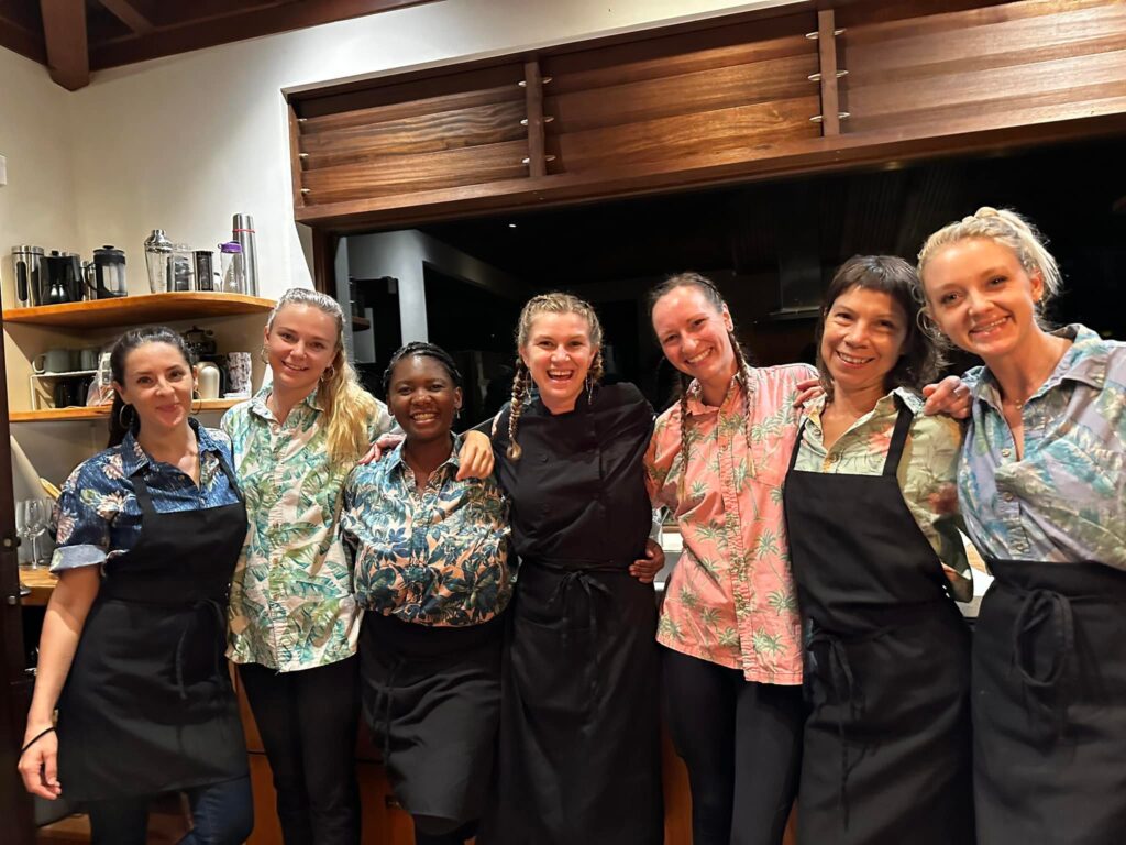 Seven women, including Chef Casia Rose, stand together in a kitchen, smiling at the camera. Most wear black aprons over colorful, patterned shirts. The background displays shelves filled with dishes and kitchen items.