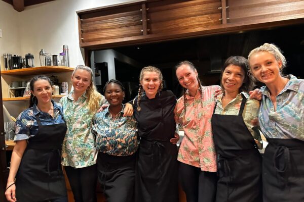 Seven women, including Chef Casia Rose, stand together in a kitchen, smiling at the camera. Most wear black aprons over colorful, patterned shirts. The background displays shelves filled with dishes and kitchen items.