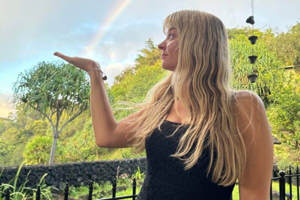 A woman with long blonde hair in a black dress stands outside, appearing to hold a rainbow on her outstretched hand, with green trees and a fence in the background—an image as vibrant as the creativity of the Lotus Chefs team.