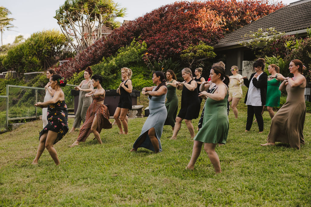 A group of women in dresses practice a hula dance on a grassy lawn near a house, surrounded by lush greenery and colorful bushes.