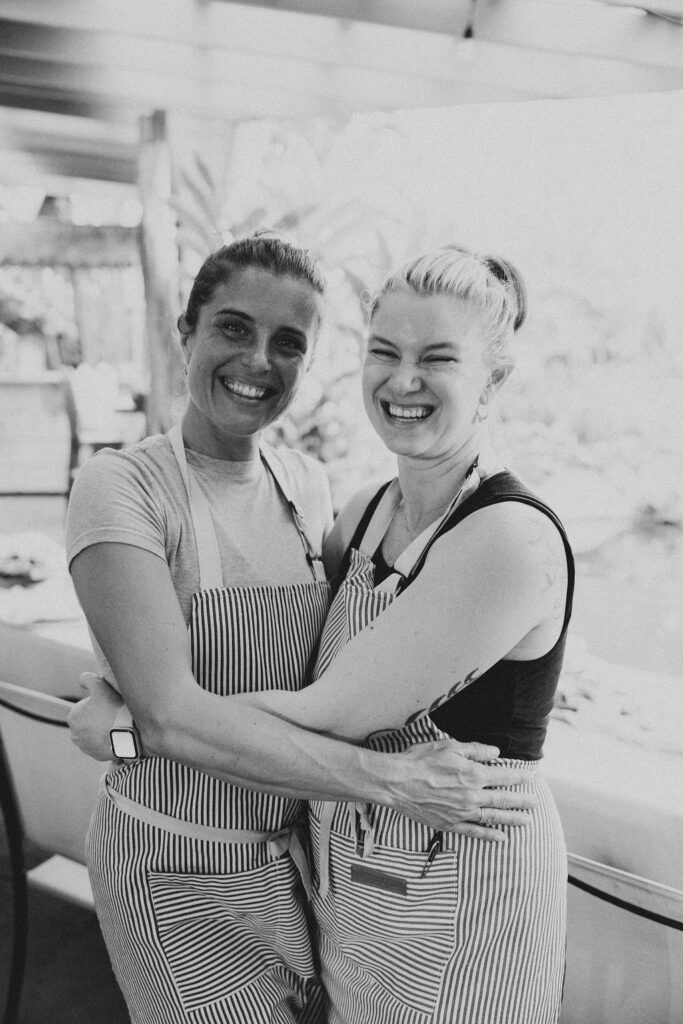Two women wearing striped aprons stand in a kitchen, smiling and hugging each other. The black and white photo captures a bright, cheerful atmosphere as Chef Angel Green shares a joyful moment with a friend.