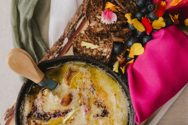 A skillet of melted cheese sits on a wooden board next to crackers, fresh berries, honeycomb, and colorful edible flowers, with a pink napkin and green and white cloth underneath.