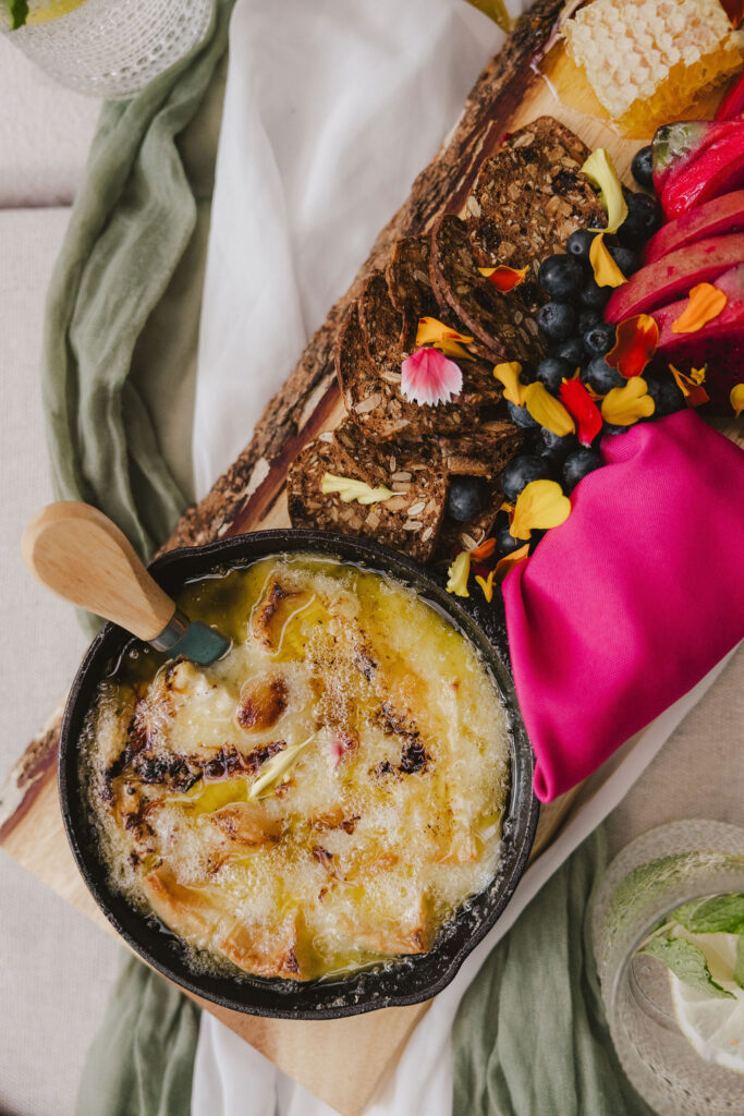 A skillet of melted cheese sits on a wooden board next to crackers, fresh berries, honeycomb, and colorful edible flowers, with a pink napkin and green and white cloth underneath.