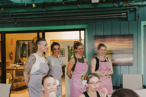 Four women wearing aprons—one of whom is Chef Angel Green—stand side by side, smiling, in front of a teal building and a large open window. People are seated in the foreground, some with flowers in their hair, near a beach painting on the wall.