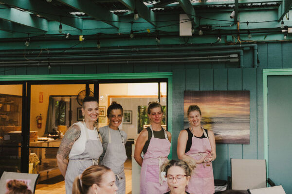 A group of people sit around a table enjoying a meal outdoors, while Chef Angel Green and three others in aprons stand and smile in the background under a covered patio with teal beams and string lights.
