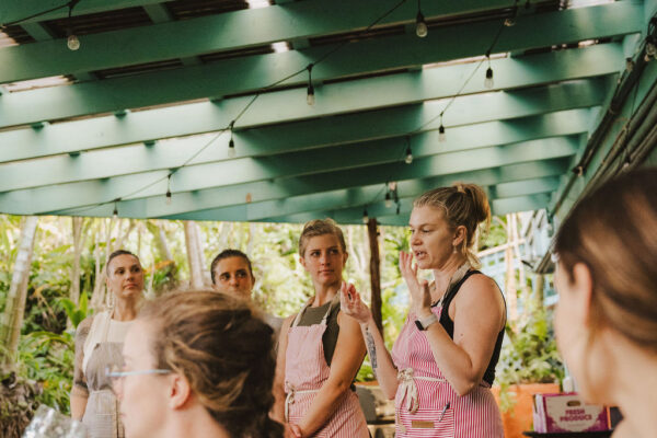 A group of women, some in pink aprons, stand and listen under a green-roofed patio decorated with string lights. Chef Angel Green speaks while others watch, and a seated woman is visible in the foreground. Lush greenery surrounds the space.