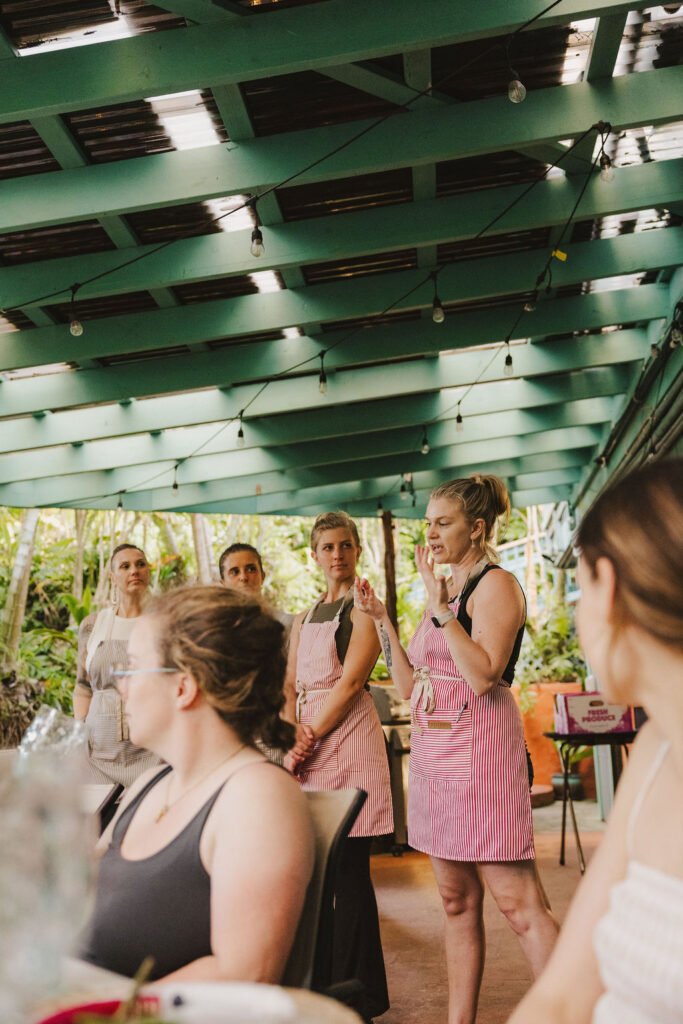 A group of women, some in pink aprons, stand and listen under a green-roofed patio decorated with string lights. Chef Angel Green speaks while others watch, and a seated woman is visible in the foreground. Lush greenery surrounds the space.