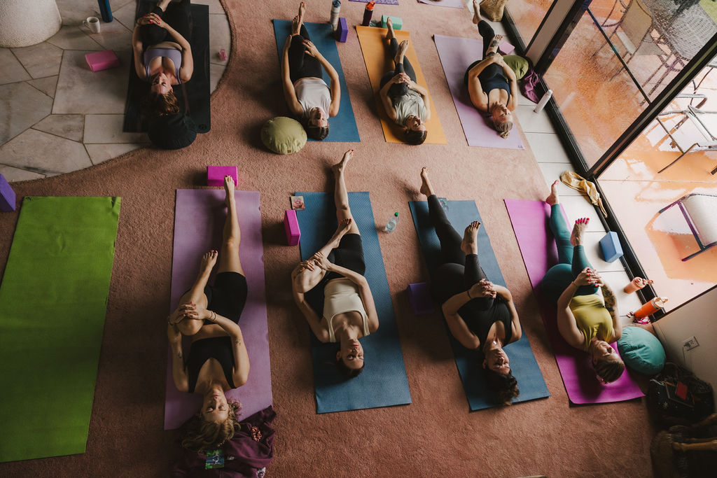 A group of people lying on yoga mats indoors, each holding one knee toward their chest, participating in a yoga class. The scene is viewed from above, showing six mats arranged in two rows.